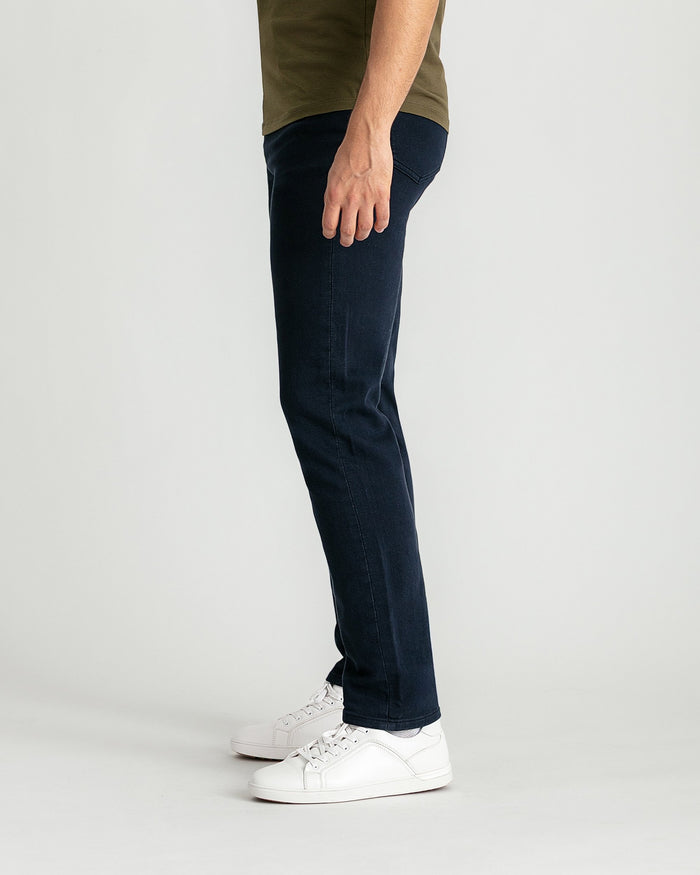 Man wearing navy slim fit jeans with olive t-shirt and white sneakers, shown from waist down in side view.