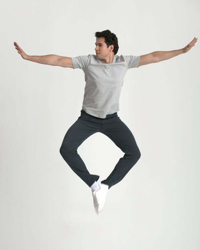 Man wearing light gray t-shirt and dark slate slim fit pants, jumping with arms outstretched.