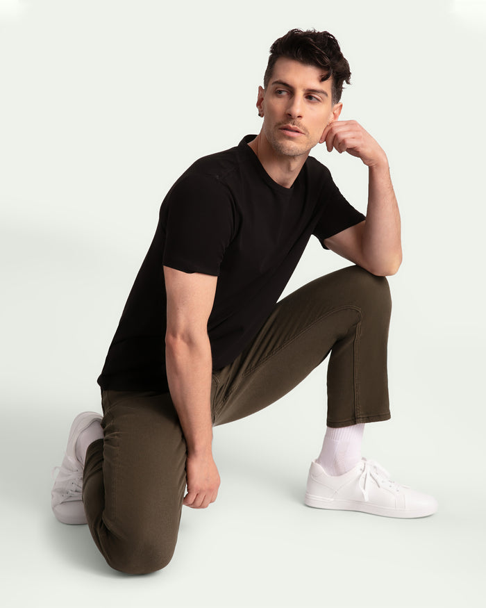 Man wearing black short-sleeve shirt and olive tapered pants with white sneakers, kneeling against white background.