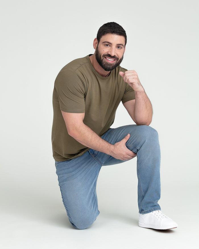 Man wearing olive short-sleeve t-shirt and light blue jeans, kneeling and posing against white background.