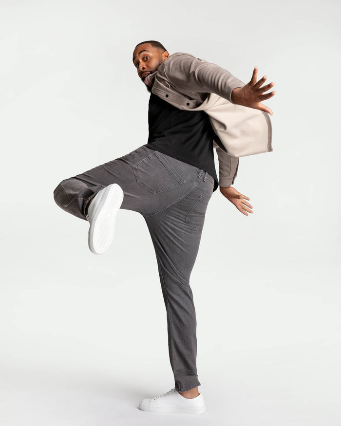 Man wearing grey slim fit jeans with black shirt, grey overshirt, and white sneakers in dynamic pose.