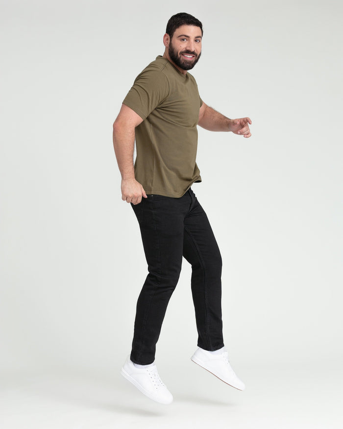 Man wearing olive short-sleeve t-shirt, black jeans, and white sneakers, jumping with a smile.