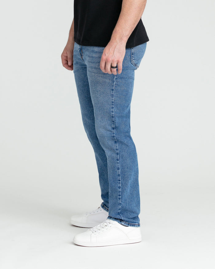 Man wearing medium blue slim-fit jeans with black shirt and white sneakers, shown from waist down in side profile.