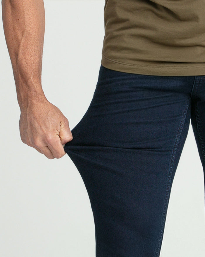 Man wearing olive-toned long-sleeve shirt and deep blue slim-fit jeans with hand in pocket.