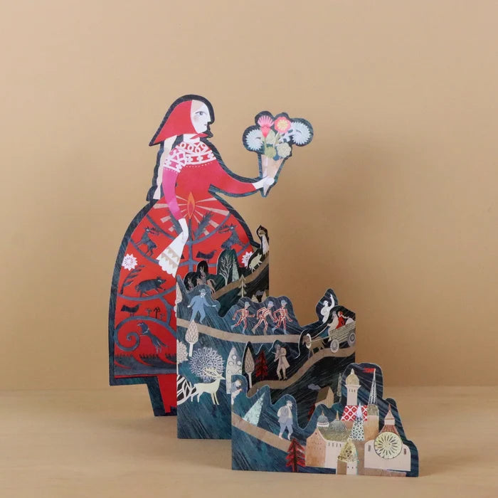 Storyteller die-cut concertina card by Sarah Young, angled view showing a tall woman in a red folkloric dress holding flowers, with layered concertina panels below depicting fairy tale scenes including figures, a sleigh, and a village, against a warm beige background