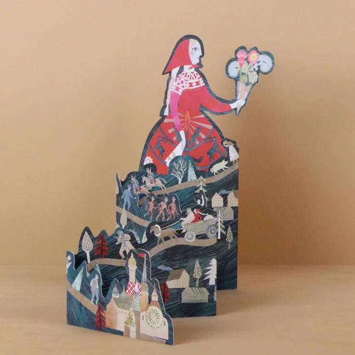 Storyteller die-cut concertina card by Sarah Young, front view fully extended to reveal a woman in a red dress holding a bouquet above layered dark teal concertina panels with fairy tale scenes including dancing skeletons, a horse-drawn cart, and a village with a church