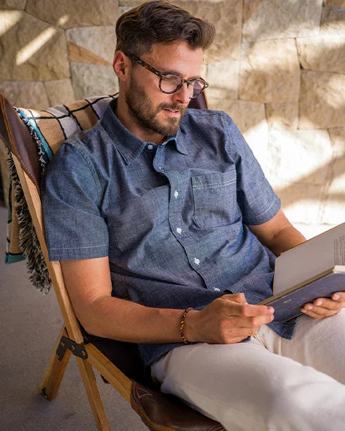 Man wearing Ace Rivington Surplus Chambray shirt in indigo rinse, seated in a wooden chair reading a book outdoors in natural light