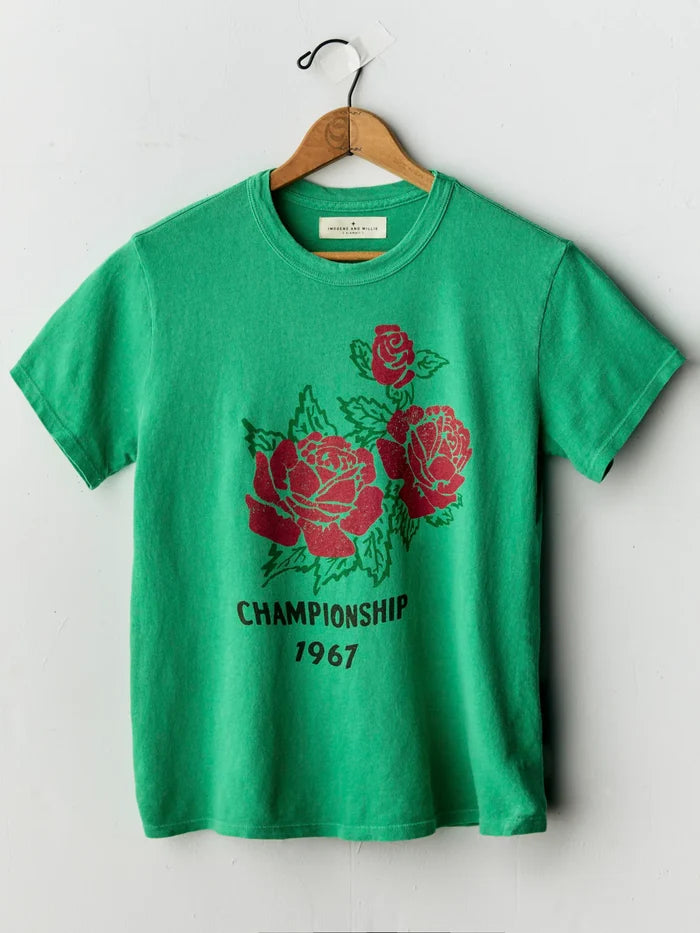 A green short-sleeve t-shirt hanging on a wooden hanger against a light gray wall. The shirt features a graphic print of three red roses with green leaves and the text 'CHAMPIONSHIP 1967' in black below the roses.