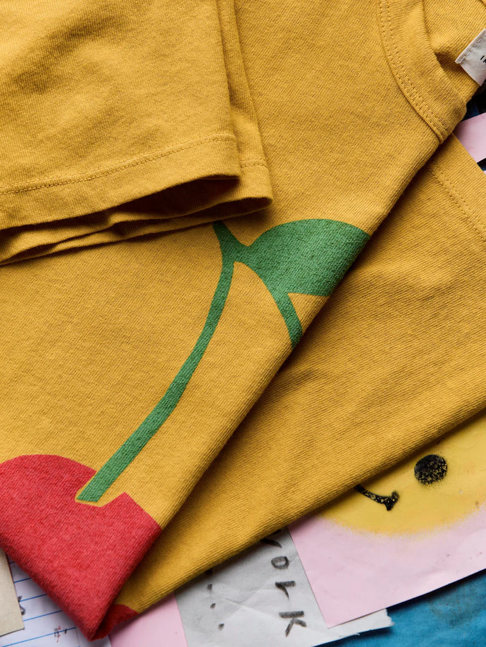Flat lay close-up of The Cherry Tee in Gold showing the screen-printed cherry stem and leaf graphic, highlighting the hand-drawn artwork detail and gold cotton fabric texture