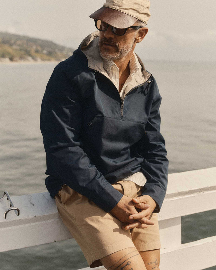 Man sitting on a boat wearing a navy jacket and beige cap with a scenic background.