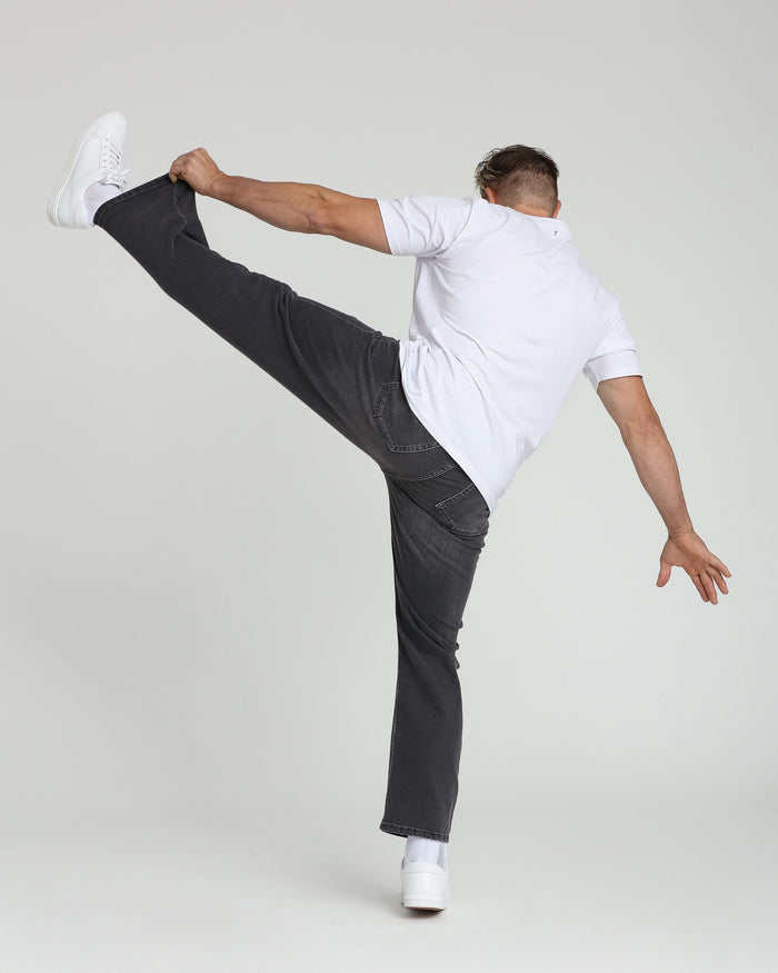Male model wearing grey relaxed-fit jeans with white sneakers and white short-sleeve shirt, posed with leg raised.