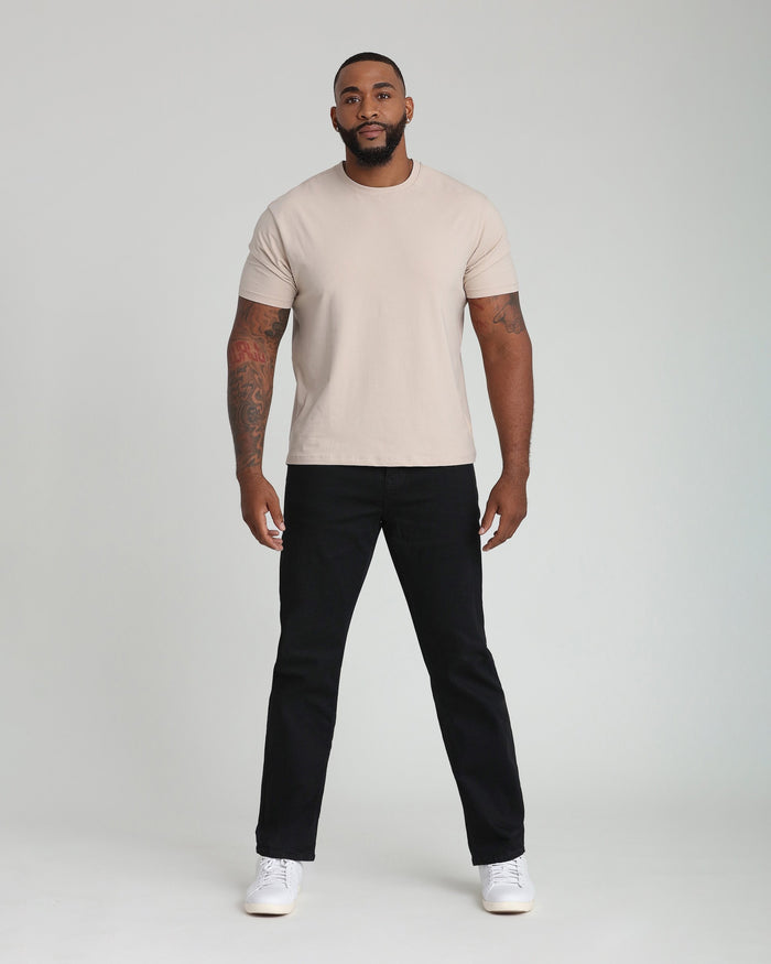 Man wearing beige short-sleeve t-shirt, black pants, and white sneakers standing against gray background.