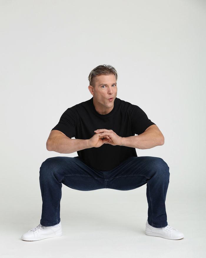 Man wearing black short-sleeve t-shirt and midnight blue jeans with white sneakers in a squatting pose.