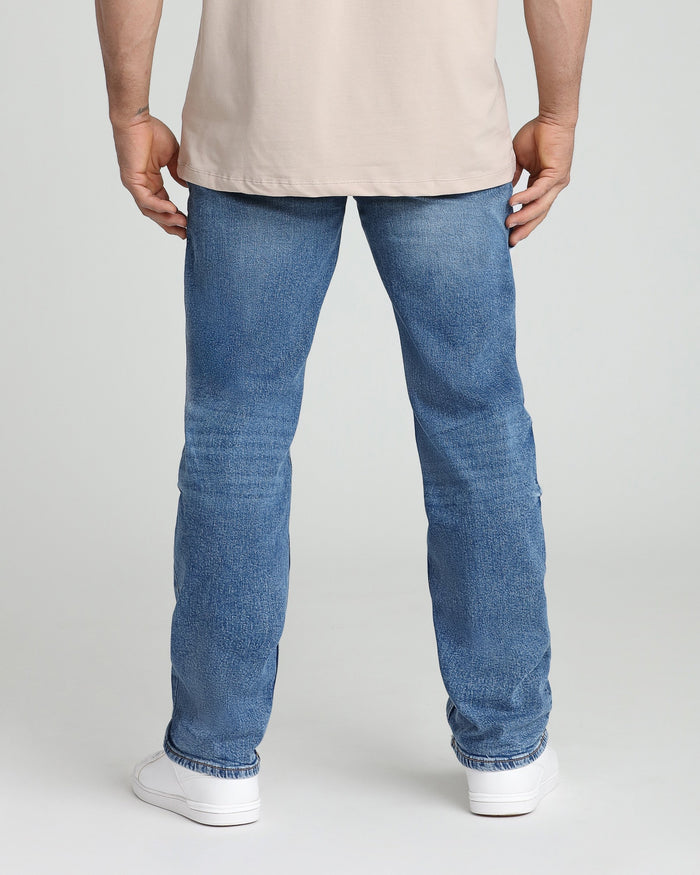 Man wearing medium blue relaxed-fit jeans with cream-colored t-shirt and white sneakers, shown from behind.