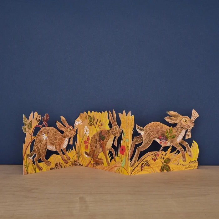 Hares Collage concertina card by Mark Hearld, fully extended showing three illustrated hares running through golden meadow grasses and wildflowers against a navy background