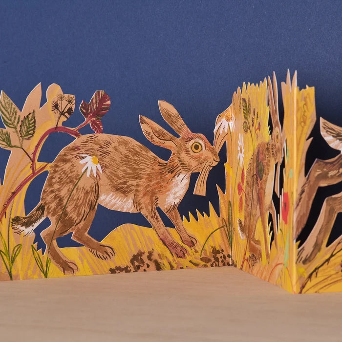 Close-up detail of the Hares Collage concertina card showing an illustrated hare leaping through golden wheat stalks and botanical foliage