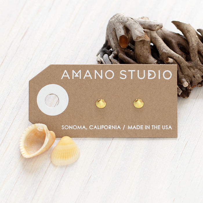 Gold shell earring studs on a brown 'Amano Studio' branded card with driftwood and seashells in the background