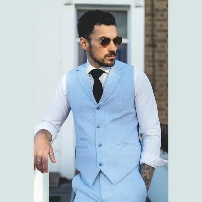 Male model wearing a stylish light blue linen summer suit with waistcoat, white shirt, black tie, sunglasses, outdoor setting