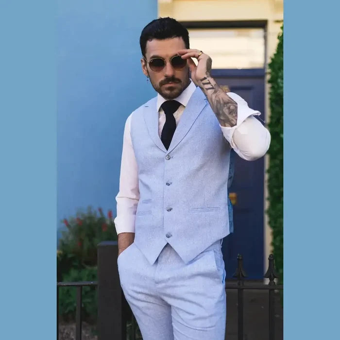 Male model wearing a light blue linen summer suit with waistcoat, white shirt, black tie, sunglasses, standing outdoors