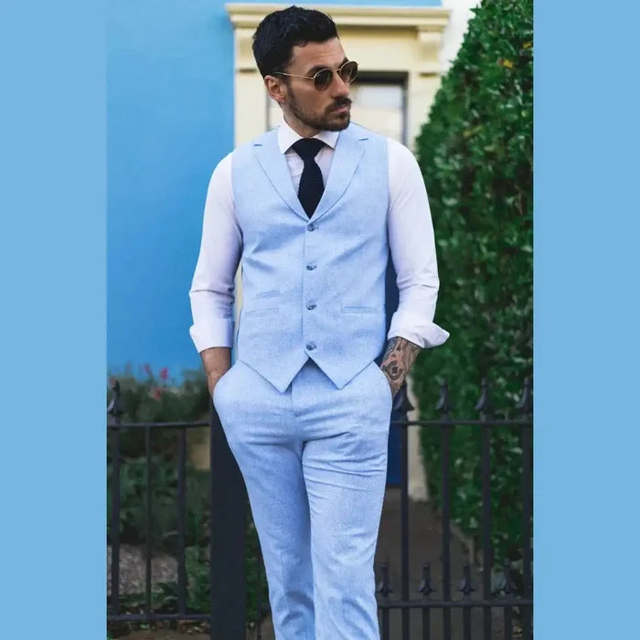 Stylish man in light blue linen suit with waistcoat and trousers, white shirt, dark tie, sunglasses, standing outdoors with greenery and blue wall background