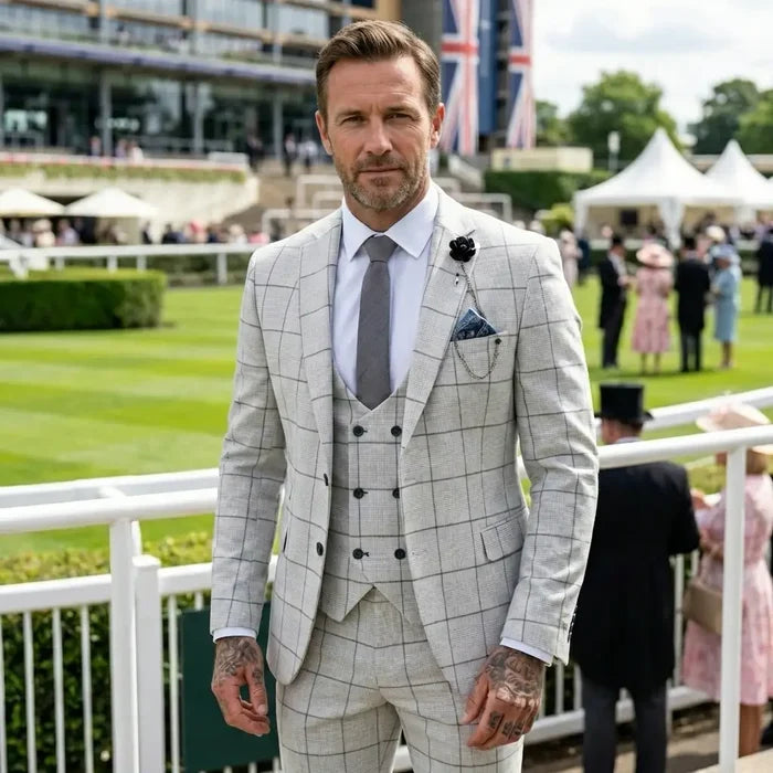 Male model wearing a stylish light grey plaid check suit with a white shirt, grey tie, and pocket square, at a racecourse event.