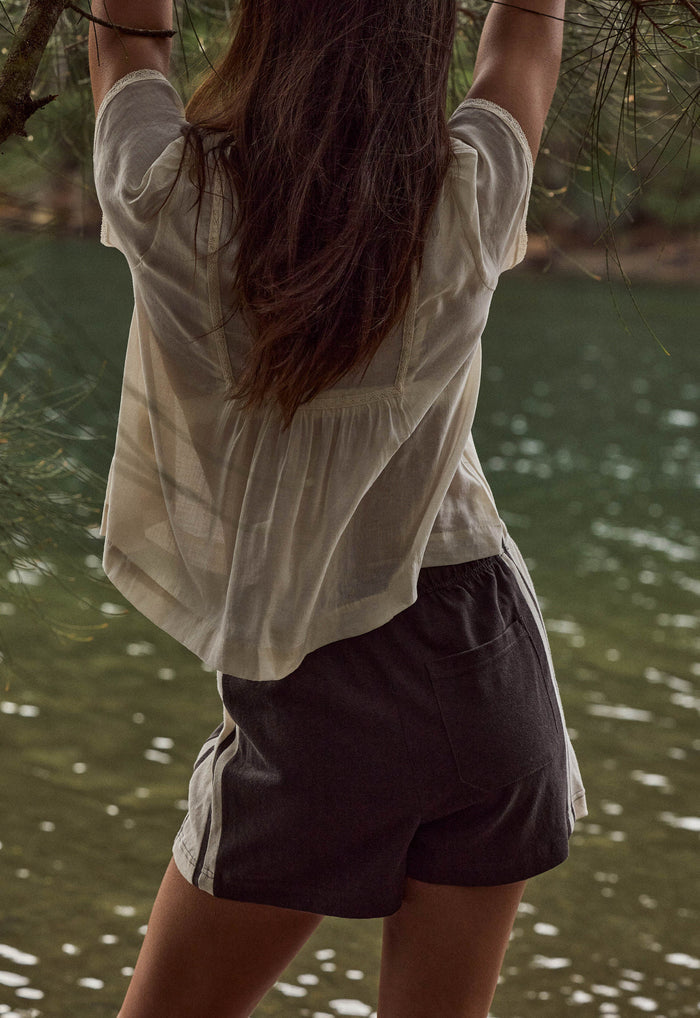 A woman viewed from behind wearing a loose off-white short-sleeve top and dark charcoal shorts with white side stripes, standing outdoors near water and hanging tree branches.