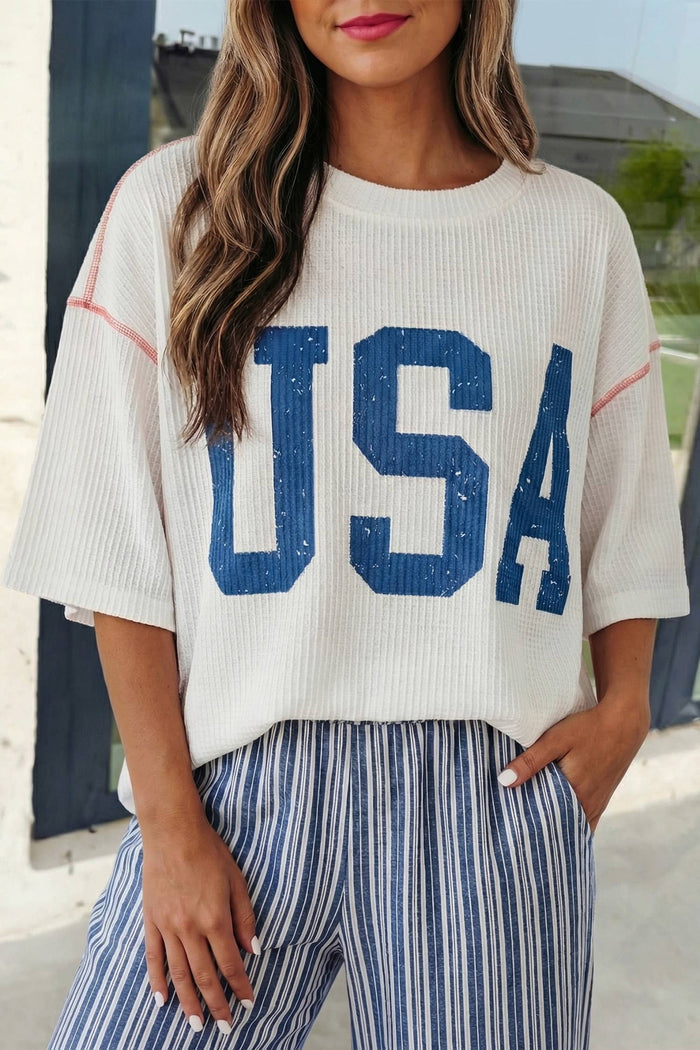 Person wearing a white sweater with 'USA' printed in blue and striped pants outdoors.