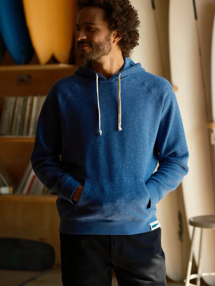 Male model wearing the Mollusk Surf Shop Whale Patch Pullover in Marine, front view showing marine blue twisted French terry hoodie with white drawstring, kangaroo pocket, and Whale Patch label on hem, photographed in a surf shop interior