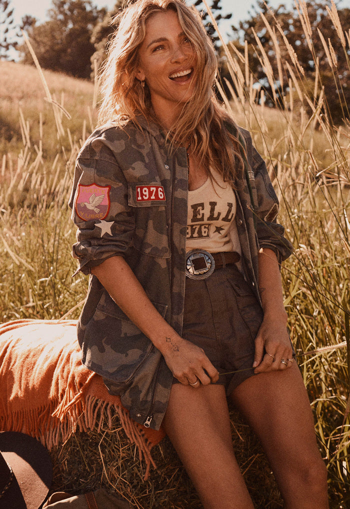 A woman sitting outdoors in a grassy field wearing a camouflage utility jacket over a beige graphic tank top and dark shorts, with a bright orange fringed blanket beside her.