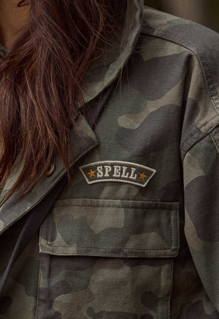 Close-up of a person wearing a camouflage utility jacket, showing the chest pocket with a sewn 'SPELL' patch on the left chest.