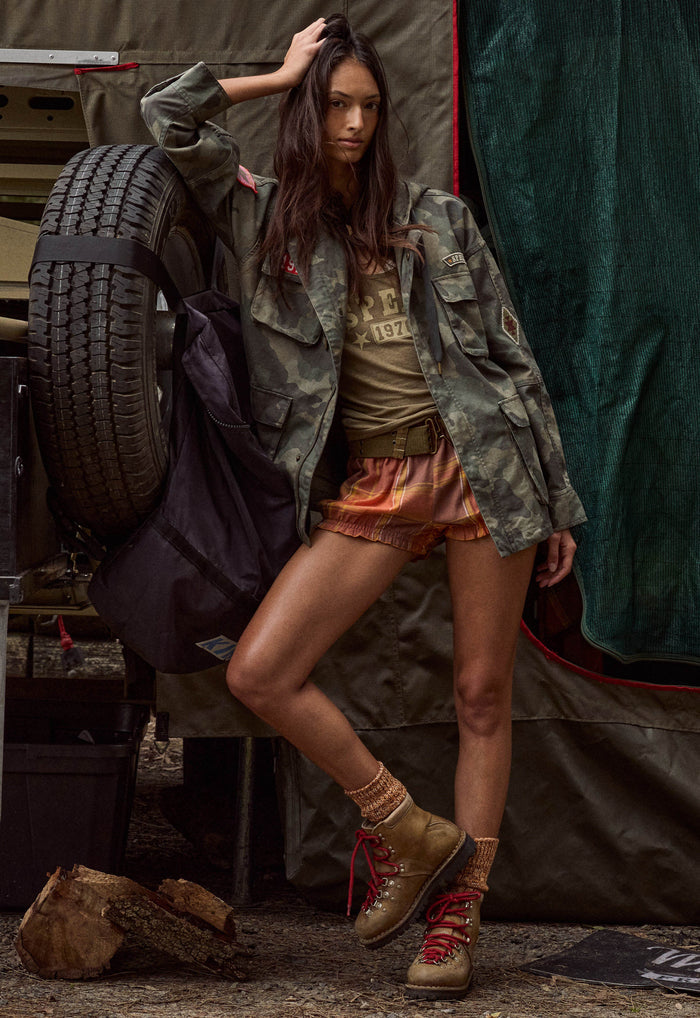 A woman poses in a rustic outdoor camping setting, wearing a camouflage utility jacket, a graphic t-shirt, orange shorts, and tan lace-up boots, with a large spare tire and tent visible in the background.