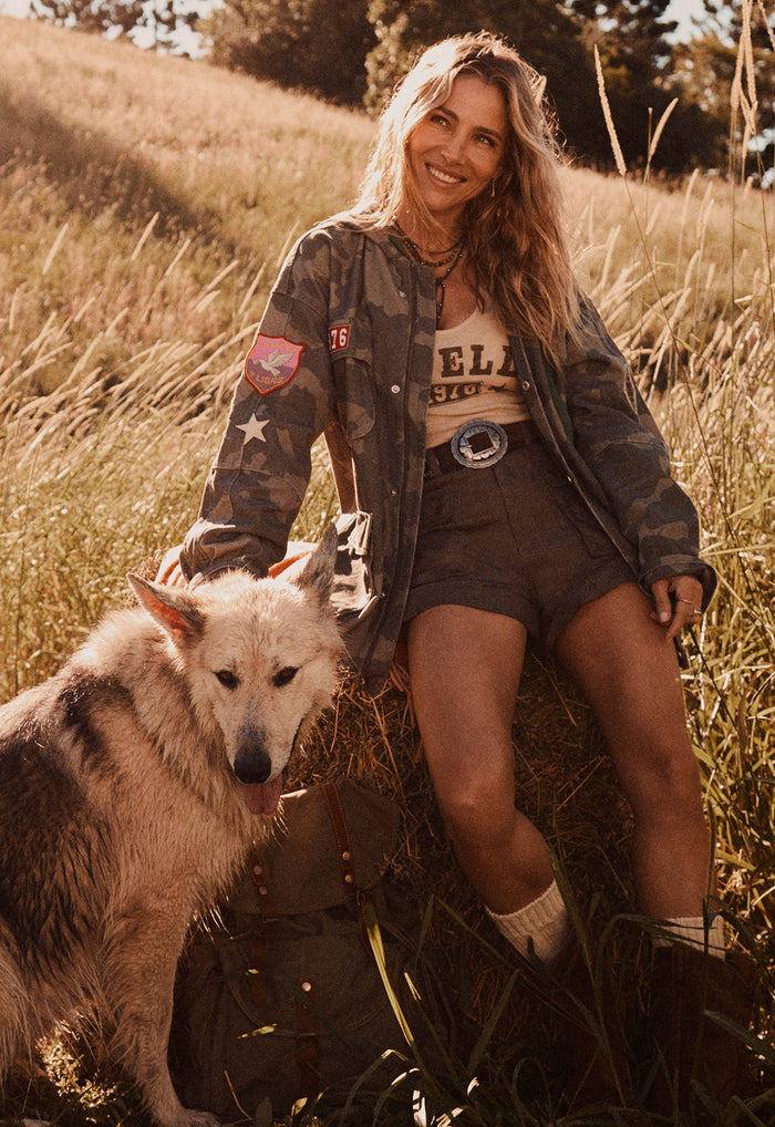 A woman sitting in a grassy field with a dog, wearing a camouflage bomber jacket over a graphic tee and shorts.