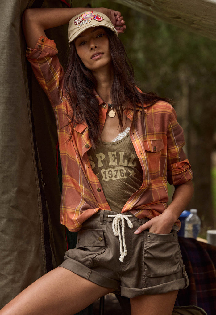 A woman posing in a rustic outdoor setting, wearing a rust-orange and yellow plaid flannel shirt over a graphic tank top, olive green cargo shorts, and a beige baseball cap.