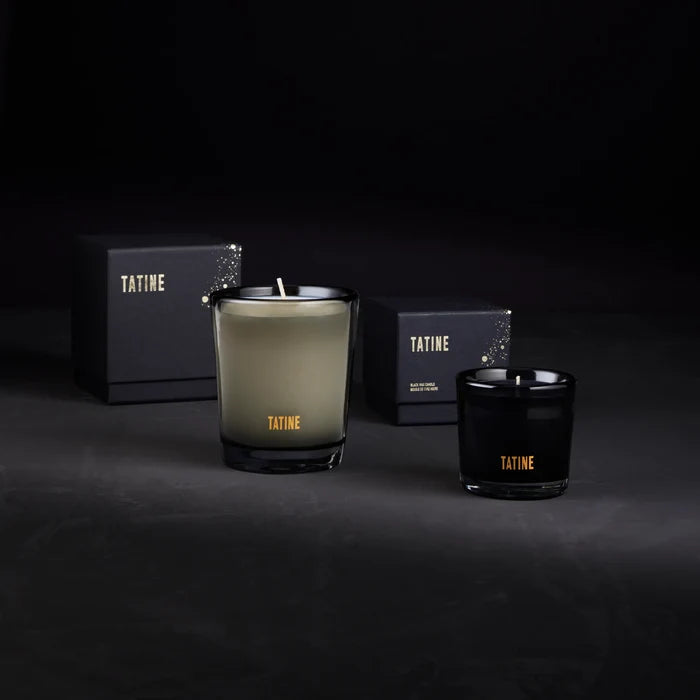 Two Tatine candles, one green and one black, with boxes in the background on a dark surface.